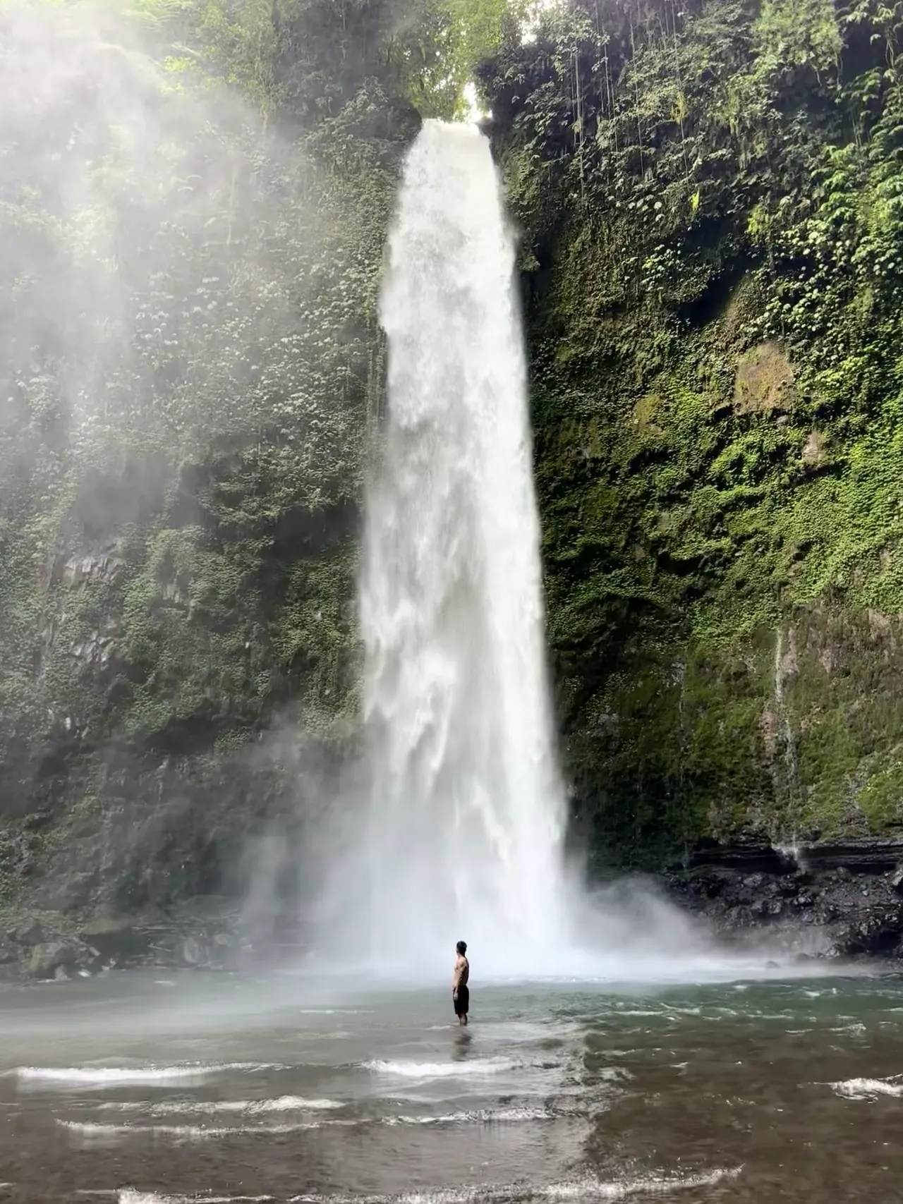 A tall waterfall in mist with a lone visitor standing in the pool